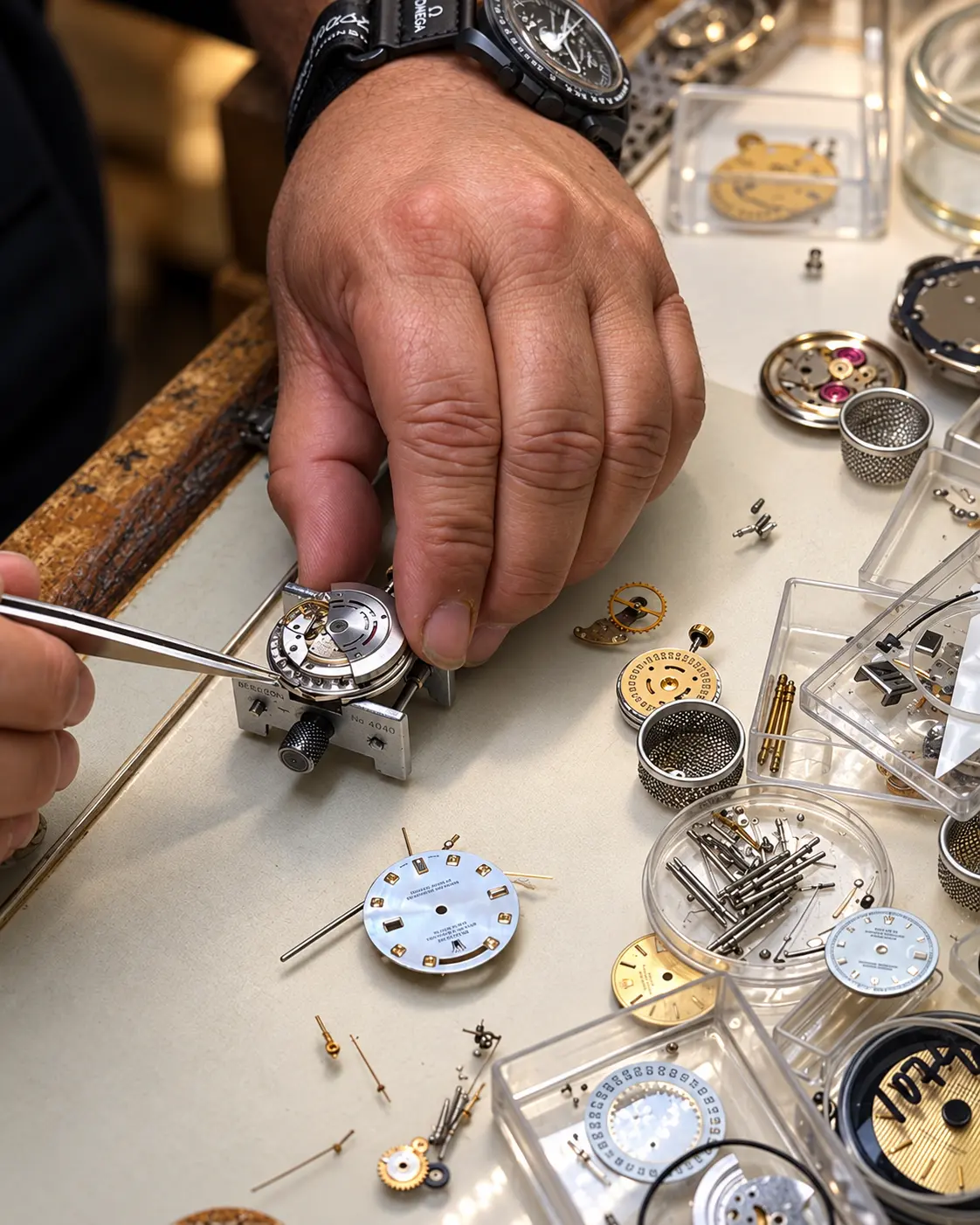 Both hands servicing a movement in a holder amid trays, parts and dials